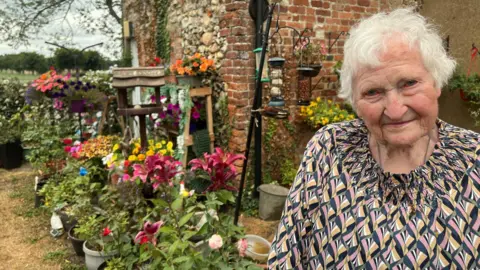 Richard Daniel/BBC Maisie Pettitt is standing in front of pots of flowers and plants, as well as bird feeders. She has white hair and is wearing a geometric patterned top.