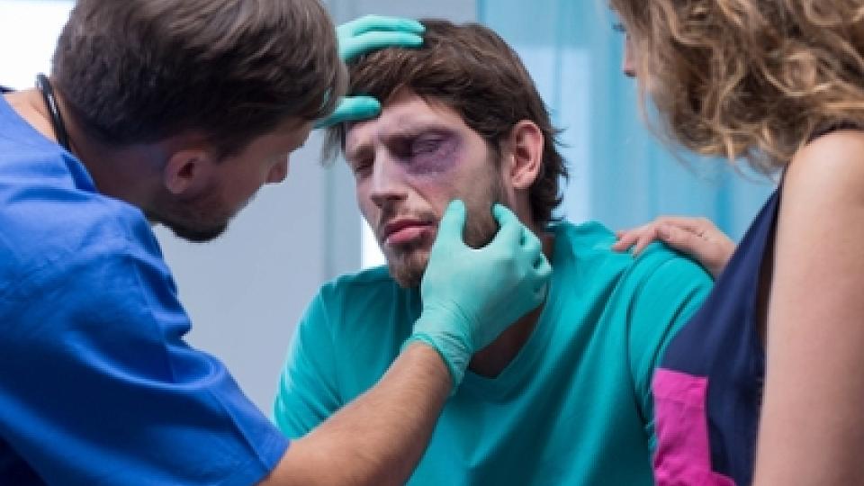 A clinician in scrubs examining a patient with facial injuries in a medical setting.