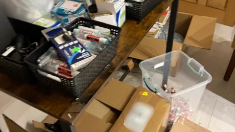 MHRA The image shows a storage room with cardboard boxes, and a clear plastic tub  with pre-filled weight-loss pens. On a table there is a black basket with more weight-loss injections