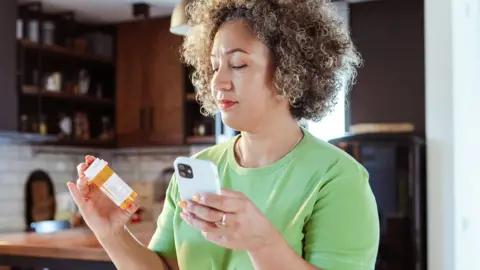 Getty Images A woman wearing a green t-shirt stands in her kitchen holding a bottle of pills in one hand and her phone in the other - she is looking at the bottle of pills