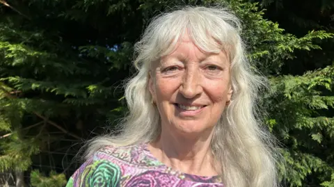 BBC Jenny Connor has long grey hair and a fringe and is wearing a top with purple and green fowers on it, smiling at the camera while standing in front of some hedgerows on a sunny day.