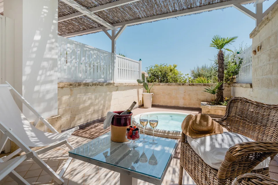 Private terrace at Canne Bianche Lifestyle Hotel in Puglia, featuring a wicker sofa with cushions, a glass table set with wine and fruit, lounge chairs, and a small jacuzzi surrounded by stone walls and tropical plants.