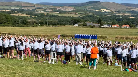 A large group of people stand gathered in a field with one arm upstretched to the side, wearing white pilates t-shirts.