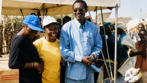 Makagbeh Princess Sesay Sesay wears a blue cap and black t-shirt and she is with a woman in a yellow t-shirt and cap and a man in a blue shirt and sunglasses. They stand at a distribution point where people are queuing.