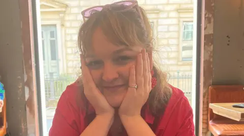 Daneka Etchells Daneka Etchells is pictured in a bright pink shirt sitting at a café table, resting her face in both hands and looking towards the camera. Large windows behind reveal a stone building outside.