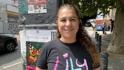 Leanne Rinne / BBC A woman, Liz Curtis, wearing a t-shirt with pink, purple and blue letters reading "Lily" on it. The dot of the letter I is a pink and purple butterfly.