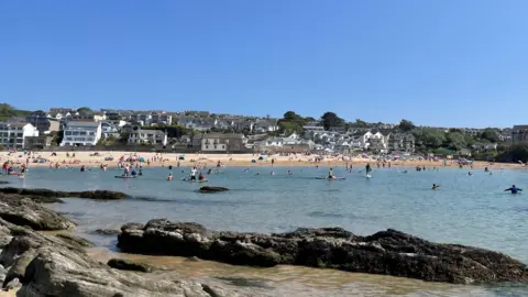 BBC People enjoying the beach at Porth near Newquay in Cornwall on a sunny day. Dozens of people are out on the water, including several on paddleboards.