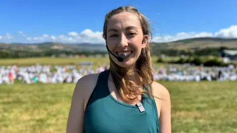 Hannah is wearing a green tank top and has a microphone mouthpiece around her ear. She is smiling in the sunshine in a field, with thousands in white t-shirts blurred behind her.