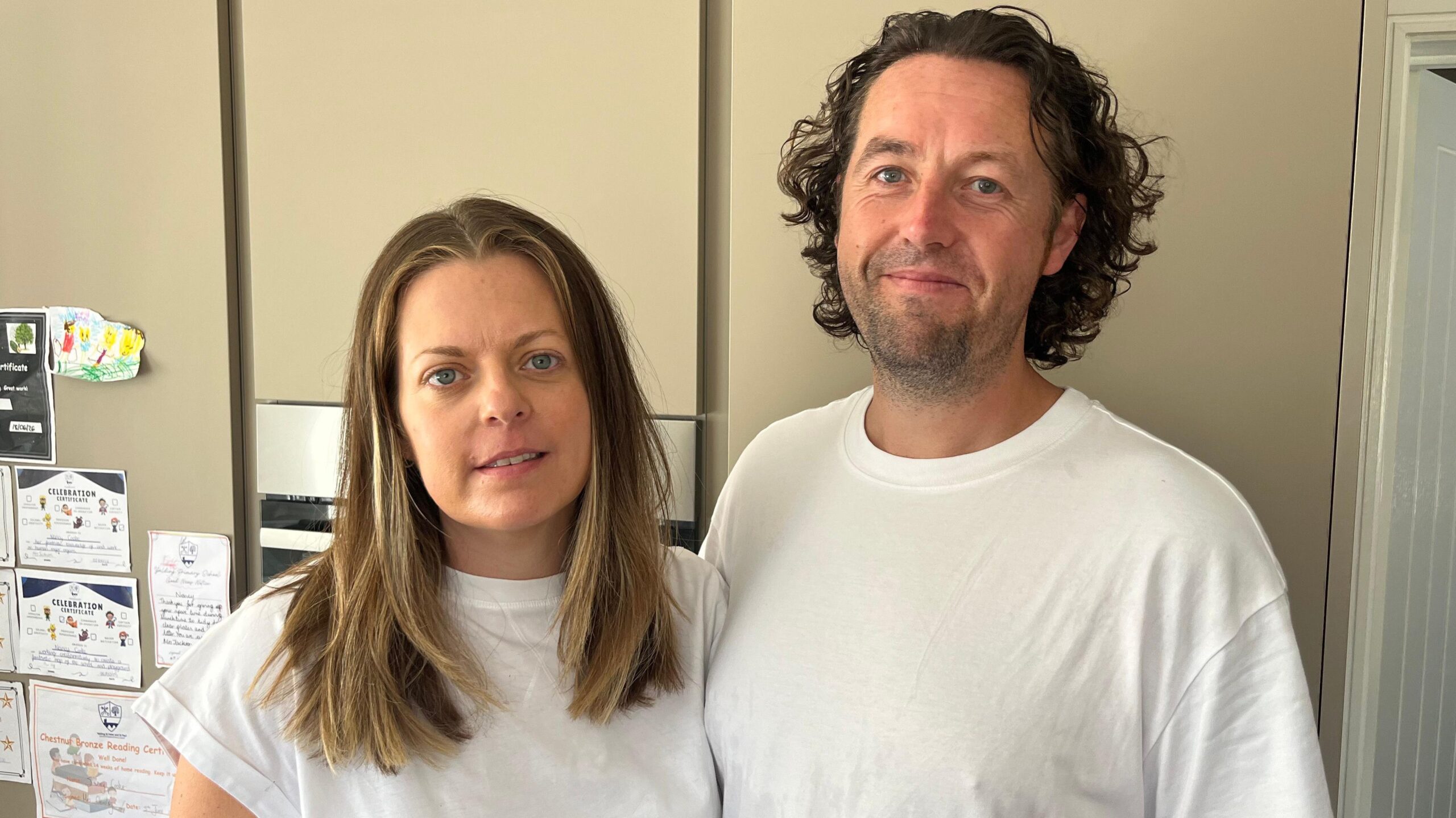A woman with short blonde hair in a white top and a man with brown hair in a curtain style in a white top pose for the camera.
