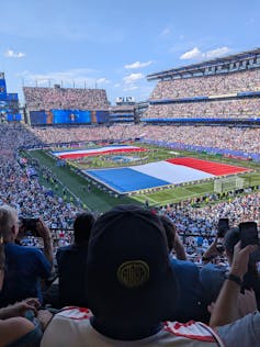 The back of a person's head, wearing a cap, in a stadium full of people while giant flags are formed on the field.