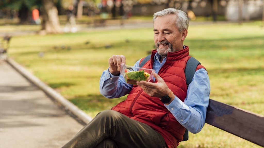 An older adult sits outside on a bench with a asalad, practicing intermittent fasting
