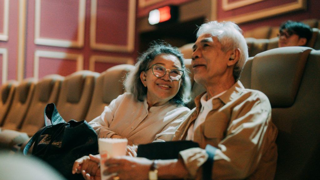 An older adult couple watches a movie at a cinema as the woman looks at her partner lovingly with a smile