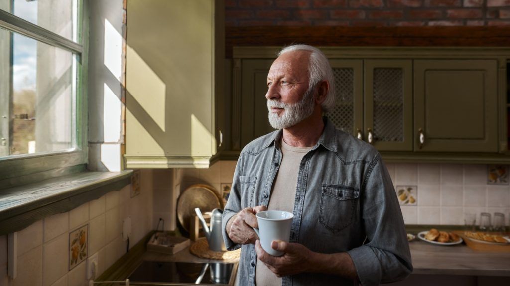 older white man holding a coffee mug while gazing out the kitchen window