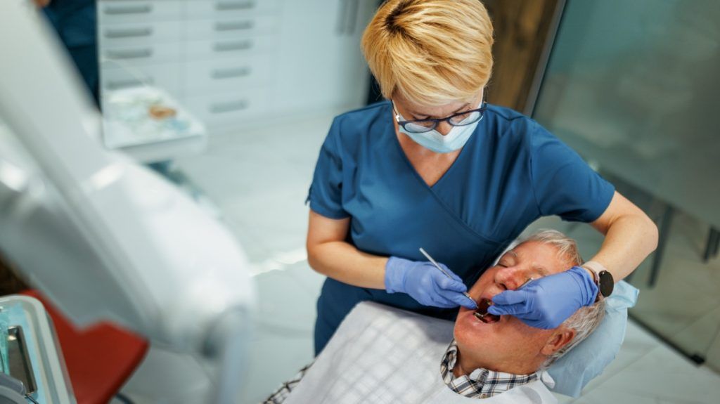 An older adult male gets his teeth checked at the dentist as a female doctor checks inside his mouth o na dentist chair