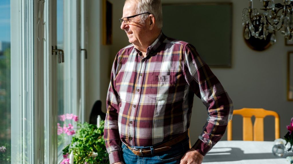 An older adult male with Parkinson's disease stands infront of a sunny window, hand on hip
