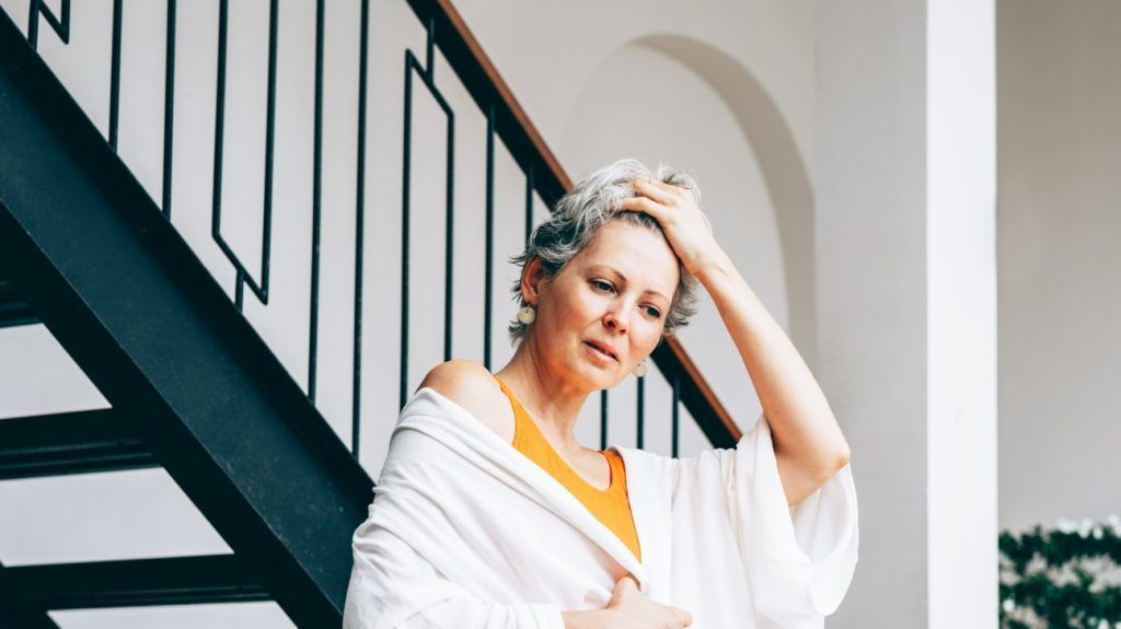Mature woman with short gray hair standing indoors near a modern staircase, wrapped in a light robe