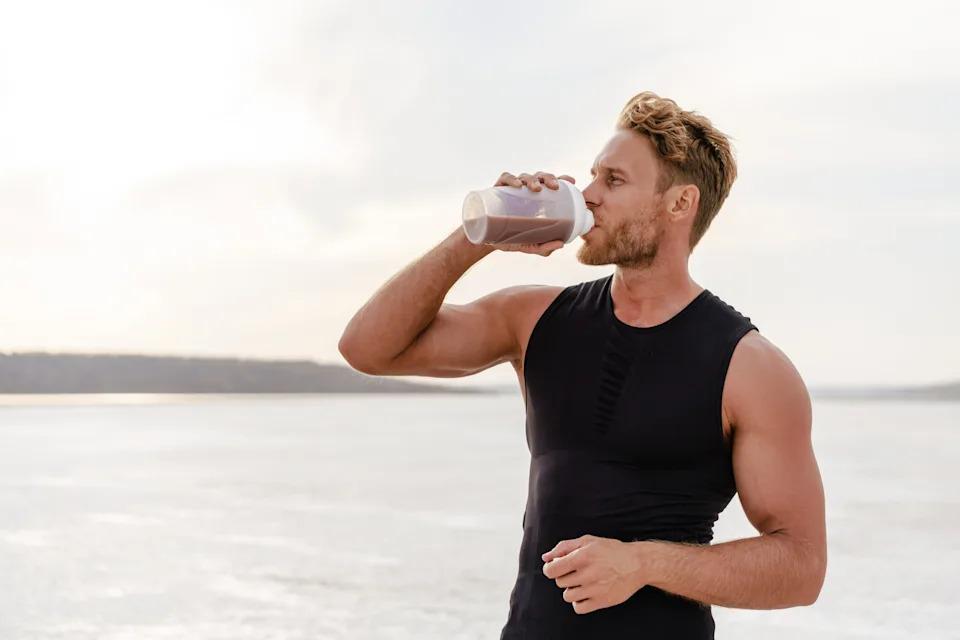 A man wearing a black athletic tank top drinks a chocolate protein shake while standing outdoors near a body of water.