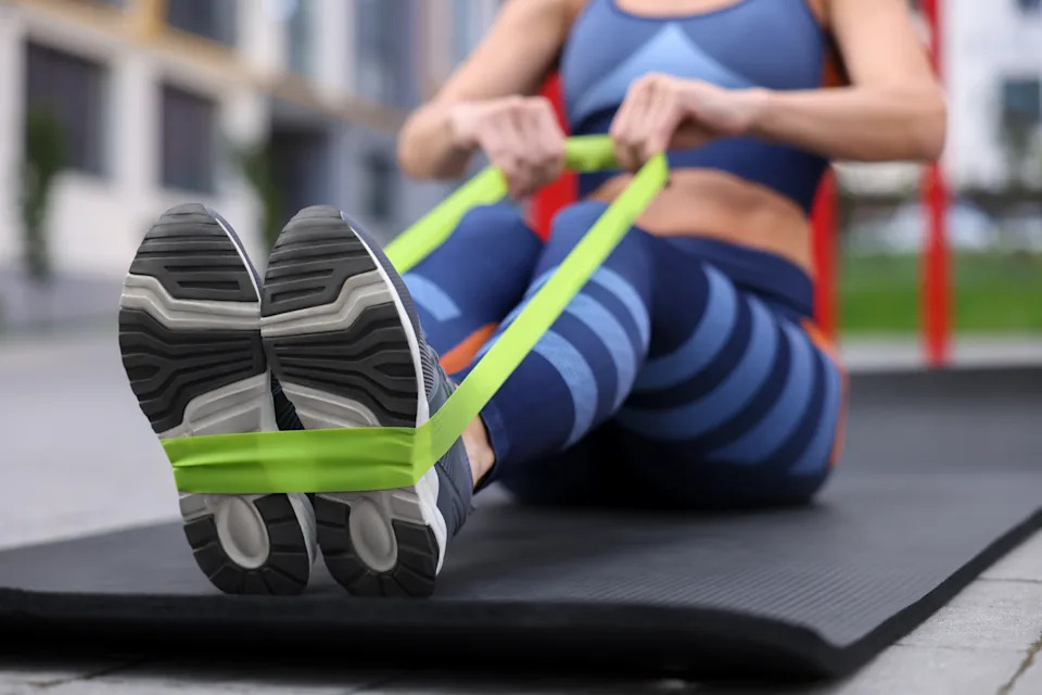 A person performs a seated rowing exercise with a green resistance band while sitting on an exercise mat outdoors.