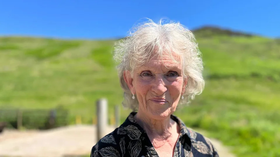 Jess Smith has short white hair and is smiling at the camera. There are green grassy hills blurred out behind her.