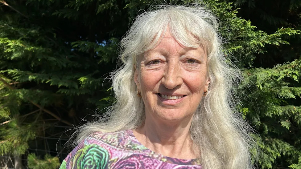 Jenny Connor has long grey hair and a fringe and is wearing a top with purple and green fowers on it, smiling at the camera while standing in front of some hedgerows on a sunny day.