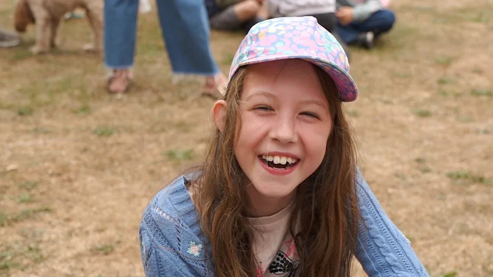 A young girl, smiling at the camera and wearing a hat with a blue jumper on. Other people are behind her, but you can only see their legs. Dried grass is behind her.