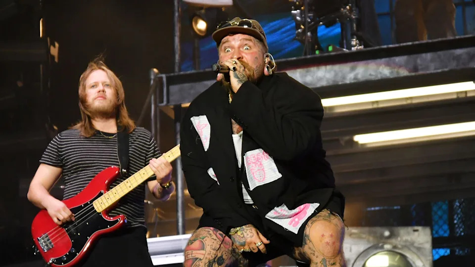 Teddy Swims performing onstage while singing into a microphone. Next to him is a bass player playing a red bass guitar.