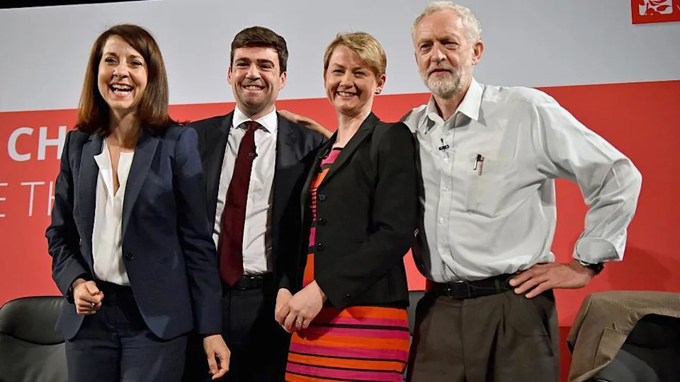 Liz Kendall, Andy Burnham, Yvette Cooper and Jeremy Corbyn. Burnham is wearing a dark suit with a white shirt and a dark red tie. Corbyn is wearing a light grey shirt with that top button undone and no tie. There are some pens tucked into this top pocket.