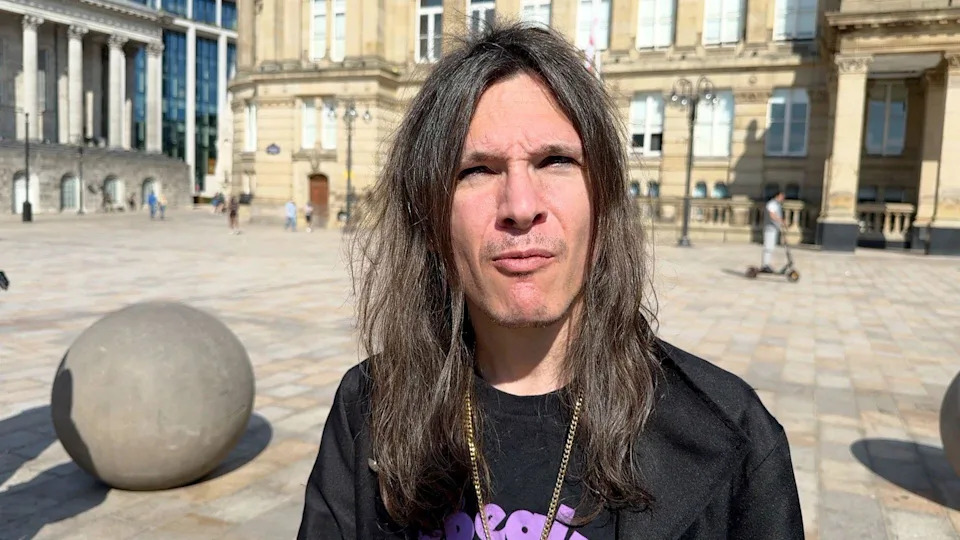 A close-up of a man with long dark hair, wearing a Black Sabbath shirt and a gold chain.