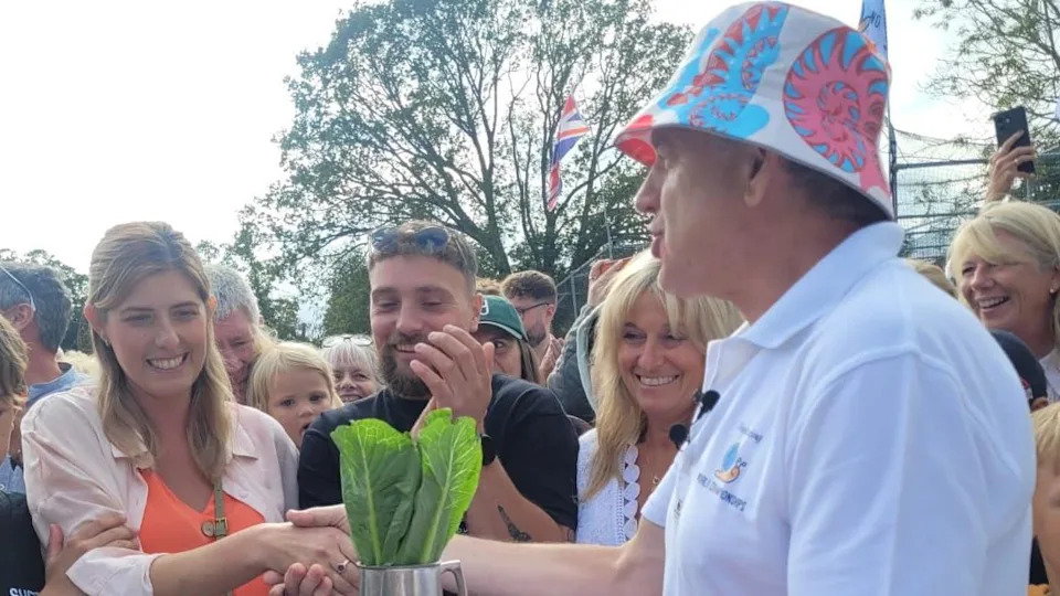 A man in a white T-shirt and a hat, shaking a woman's hand, holding a tankard with lettuce in. Other people are standing around.