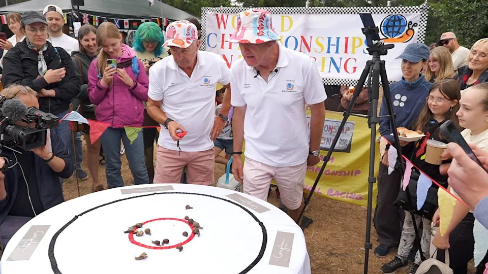 A large group of people standing around a white table, with a cloth and snails on it. Two men have white T-shirts and hats on. A camera stand is to the right.