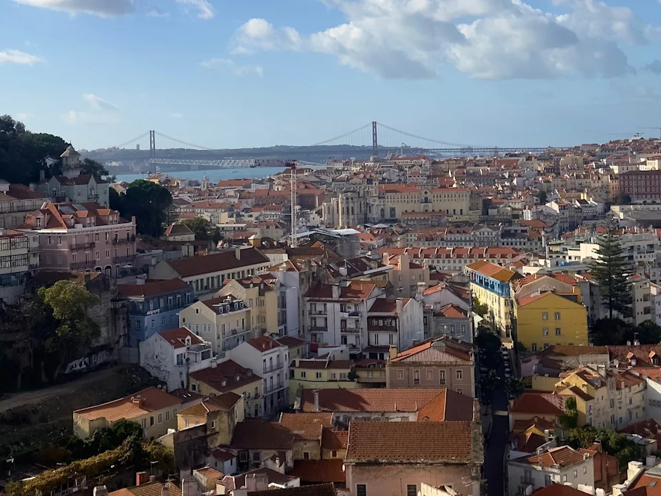 Lisbon rooftops stretch toward the 25 de Abril Bridge under a partly cloudy blue sky.