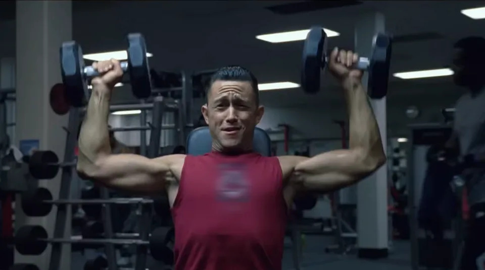 A person in a gym is lifting weights while seated, appearing focused and exerting effort. Exercise equipment is visible in the background