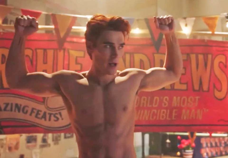 A shirtless man with toned muscles raises his arms in front of a carnival-themed banner