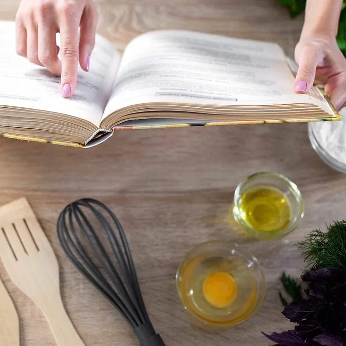 A hand points at a recipe in an open cookbook, surrounded by kitchen tools, bowls of oil, an egg, and fresh herbs on a wooden surface.