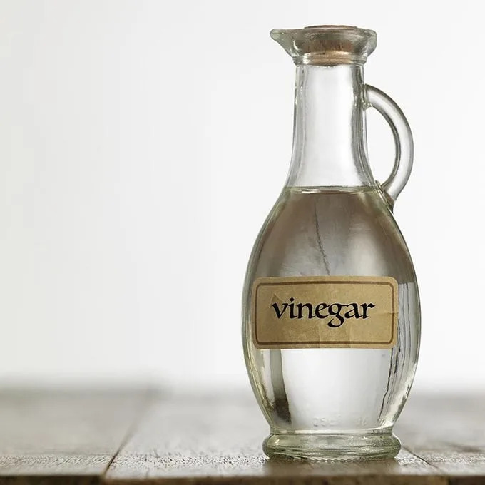 A glass vinegar bottle stands upright on a wooden surface, filled with clear liquid, against a soft, neutral background.
