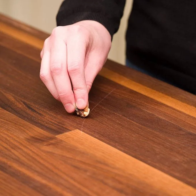A hand rubs a piece of food across a scratched wooden table surface, attempting to clean or repair the damage in a home kitchen environment.