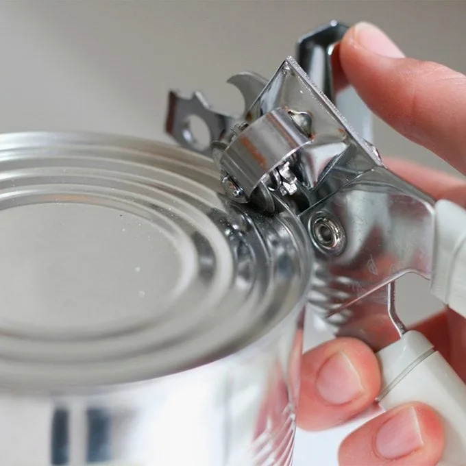 A hand holds a can opener against a metal can's lid, turning the lever to pierce the surface, preparing to open it in a kitchen setting.