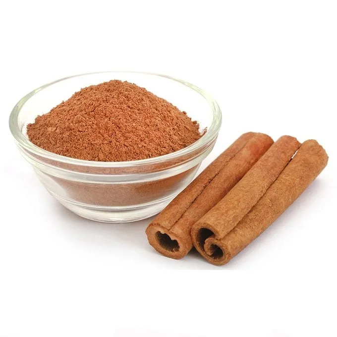 A bowl of ground cinnamon rests beside several cinnamon sticks, displaying a rich brown color against a plain white background.