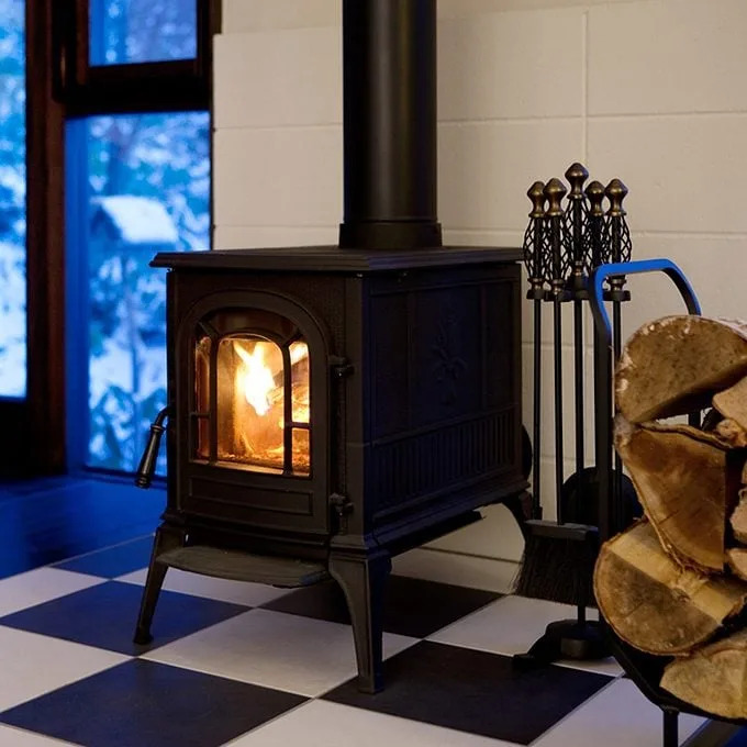 A black wood stove emits a warm glow, surrounded by a checkerboard floor and firewood, next to a window revealing a snowy landscape outside.
