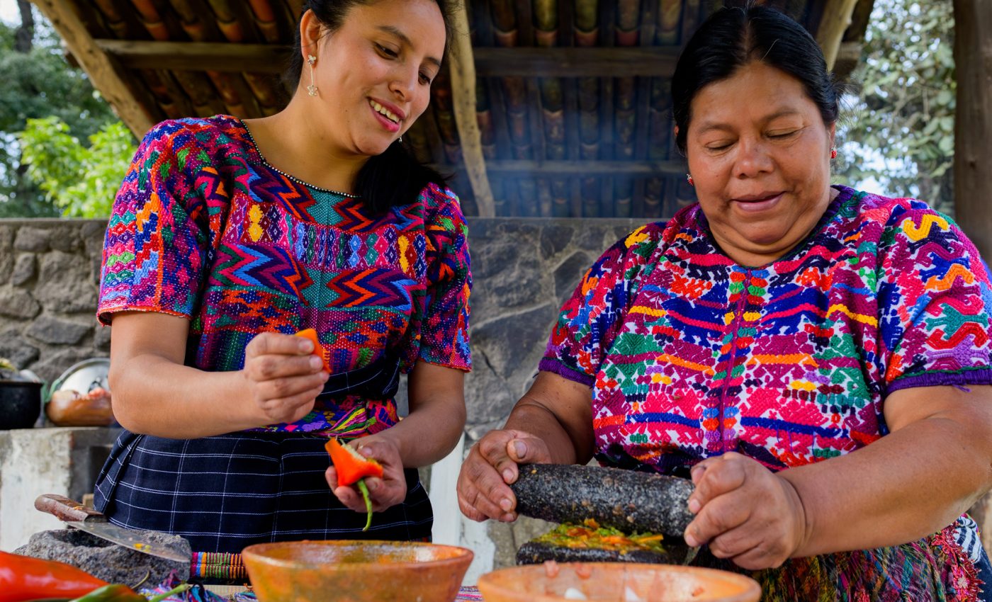 Indigenous food knowledge shared as two Mayan women prepare traditional food using a stone grinding tool in Guatemala.
