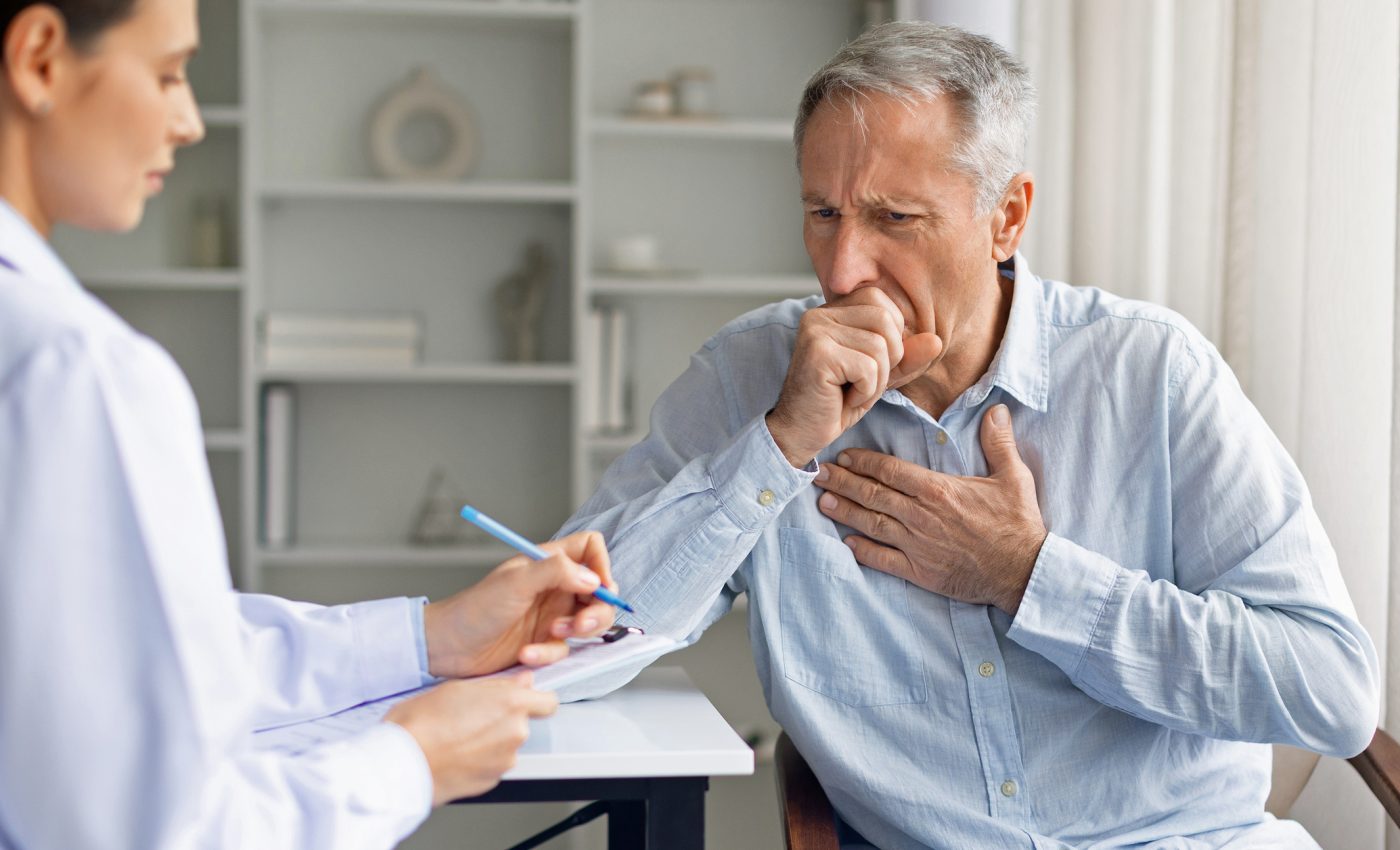 Older man coughing during a medical appointment as a doctor takes notes, illustrating how longer lifespans are leaving people with more years of poor health.