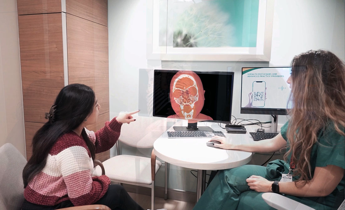 Person points to a computer screen displaying a 3D skull and sinus image during a clinical consultation, while a clinician in scrubs sits beside them at a desk with dual monitors in a medical office.