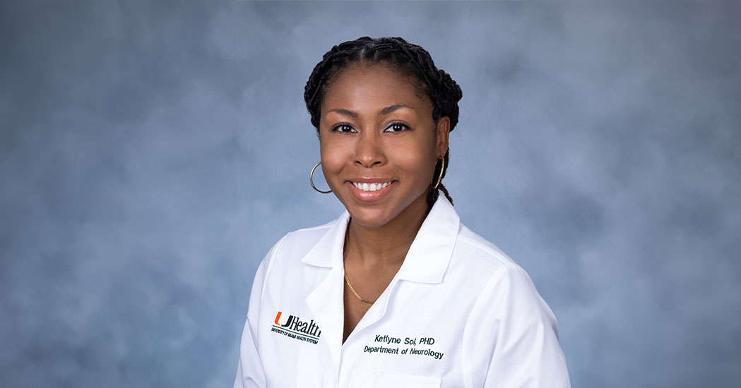 Portrait of Ketlyne Sol, Ph.D., research assistant professor of cognitive neurology at the University of Miami Miller School of Medicine, wearing a white clinical coat against a blue studio background.