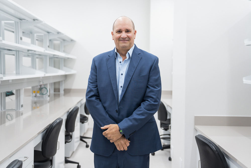 Dr. Leonard Petrucelli in a blue blazer and checkered shirt stands in a bright laboratory space with empty workbenches and chairs behind him.