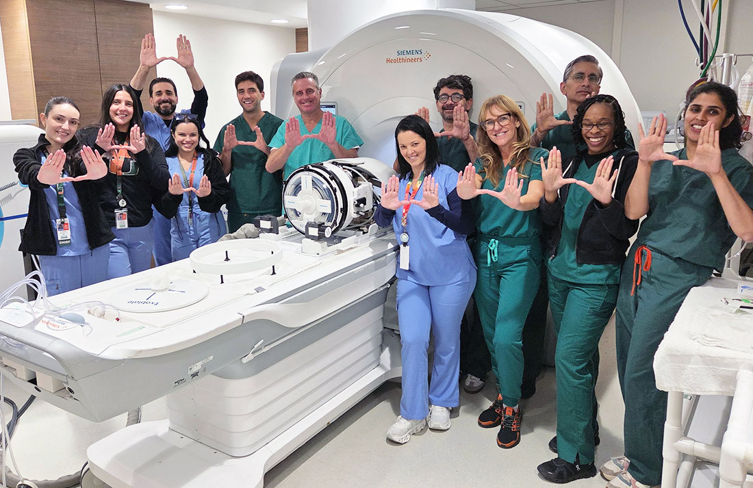 Clinical team members at UHealth stand beside an MRI-guided focused ultrasound system, arranged around the scanner table and holding their hands in a “U” shape inside a hospital procedure room.