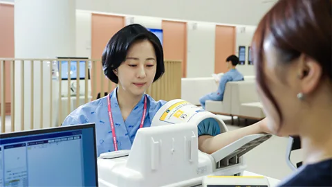 KMI Medical Institute A woman in a blue medical gown inserts her arm into a blood pressure cuff (Credit: KMI Medical Institute)