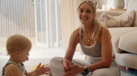Family handout Becki Barratt is sitting on the floor in her living room next to her baby daughter Aréya, who is clapping her hands.