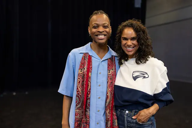 A thin, Black man in his early 20s wearing a button-down blue shirt and a red scarf stands beside a Black women in her early 40s with curly black, shoulder-length hair. Both wear broad smiles.