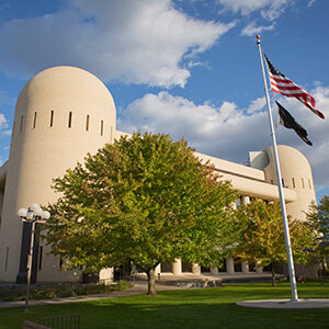 Spokane Regional Health District's main campus at 1101 West College Avenue in Spokane, Washington. The building is shown with a blue sky behind it. Maple trees in front of the building are mostly green, touched with red. A flag pole in front of the building displays the American flag and the National League of Families Prisoners of Warm Missing in Action flag.