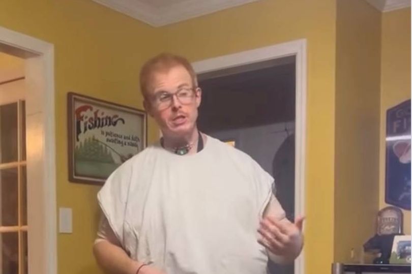 An individual dressed in a white garment is standing in a room with yellow walls and gesturing with his hands, perhaps explaining or demonstrating something. The room is adorned with a framed picture and other decorative items.